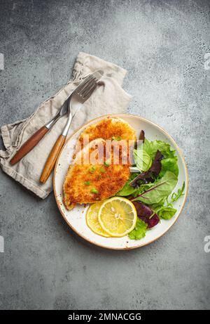 Knuspriges paniertes Hähnchenfilet mit grünem Salat und Zitrone auf dem Teller mit Besteck auf grauem rustikalem Betontisch von oben Stockfoto