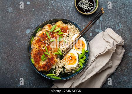 Asiatische Nudeln Ramen-Suppe mit frittiertem Panko-Hühnerfilet und gekochten Eiern in einer Keramikschüssel mit Hackstangen und Sojasoße auf rustikalem Steinhintergrund Stockfoto