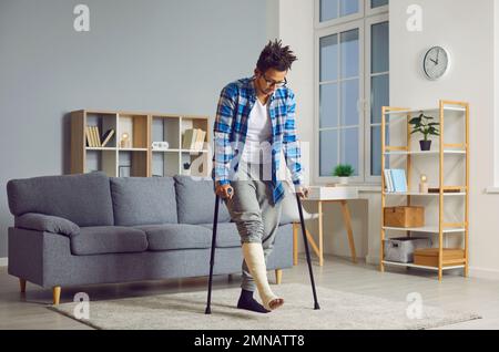Junger afroamerikanischer Mann mit gebrochenem Bein in einem Gips, der zu Hause mit Krücken läuft Stockfoto