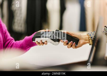 Nahaufnahme von der Seite einer nicht wiedererkennbaren Frau, die per Smartphone NFC in der Boutique für Kleidung und in Kopierbereichen bezahlt Stockfoto