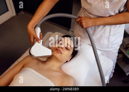 Junge brünette Frau, die beim Besuch des Schönheitszentrums im Salon eine Fotoepilierung erhält, professionelle Kosmetikerin, die Laserhaarentfernung durchführt Stockfoto