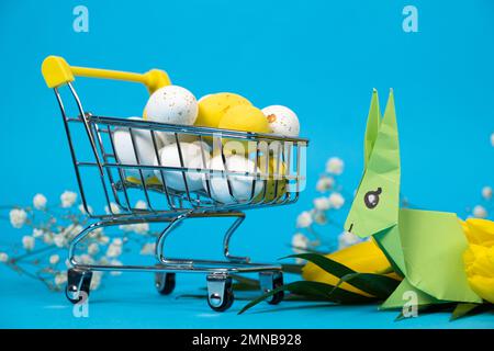 Osterweiß und -Gelb bemalte Eier und Häschen in einem Einkaufswagen auf blauem Hintergrund. Osterangebote und -Shopping Stockfoto