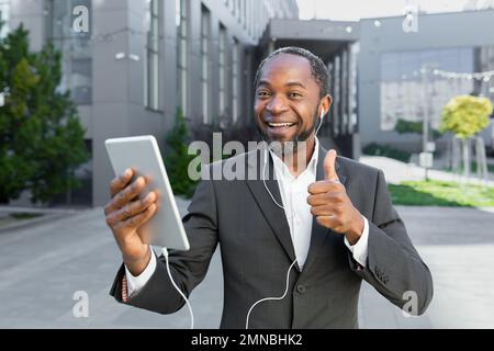 Ein junger afroamerikanischer Mann steht draußen in der Nähe eines Bürozentrums, trägt Kopfhörer und spricht bei einem Videoanruf. Hält ein Tablet in seinen Händen, zeigt einen Superhelden, lächelt, schaut in die Kamera. Stockfoto
