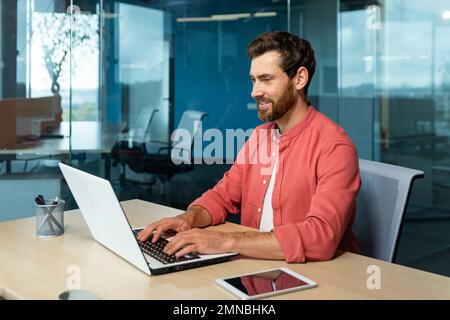 Fernunterricht. Porträt eines jungen männlichen Schülers in einem roten Hemd. Am Schreibtisch im Büro sitzen und auf einem Laptop online lernen. Schreibt ein Diplom, einen Kurs, einen Abstrakt, einen Vortrag. Stockfoto