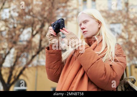 Junge Albino-Frau in warmbrauner Jacke und Schal mit Fotokamera, während sie auf eigene Faust Fotos von der Herbststadt macht Stockfoto