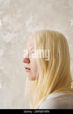 Junge, ruhige Albino-Frau mit glattem, langem Haar, die nach vorne blickt, während sie isoliert vor der Kamera über einer pastellfarbenen Wand steht Stockfoto