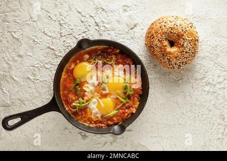 Shakshuka in einer gusseisernen Pfanne mit frisch gebackenem Bagel auf weißem, strukturiertem Hintergrund. Spiegeleier mit Tomatensoße. Shakshuka, eine traditionelle Mahlzeit des je Stockfoto