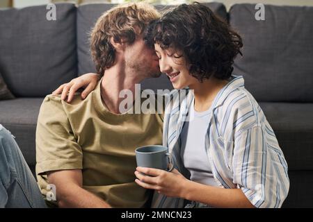 Junges interkulturelles romantisches Paar, das sich auf der Couch im Wohnzimmer erholt, während eine hübsche Frau mit einer Tasse Kaffee ihren Mann umarmt Stockfoto