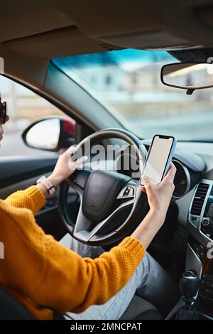 Hände einer unbekannten Fahrerin, die während der Fahrt ein Mobiltelefon mit weißem Bildschirm verwendet Stockfoto