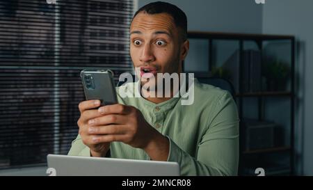 Aufgeregter afroamerikanischer Geschäftsmann, der E-Mails erhielt und gute Nachrichten auf dem Handy las, überglücklich Mann Gewinner Unternehmer, der im Büro saß und feierte Stockfoto