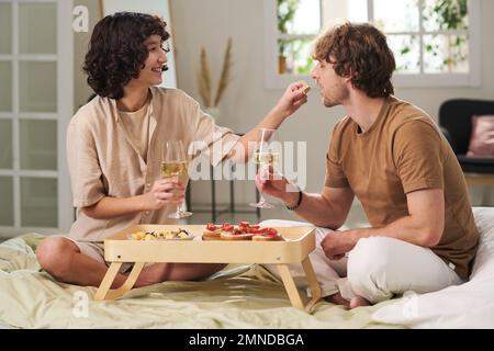 Glückliche junge Frau mit einer Flöte Champagner, die ihrem Mann ein Stück Snack in den Mund steckte, während beide das Frühstück im Bett genossen Stockfoto