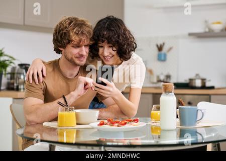 Junge lächelnde Frau, die ihrem Mann auf dem Smartphone etwas zeigt, sitzt am Tisch mit Sandwiches, Saft und Milch und frühstückt Stockfoto