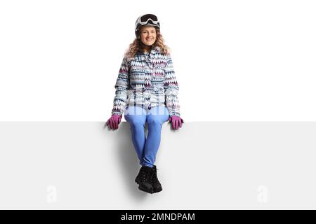 Junge Frau mit Skiausrüstung, die auf einer leeren Platte sitzt, isoliert auf weißem Hintergrund