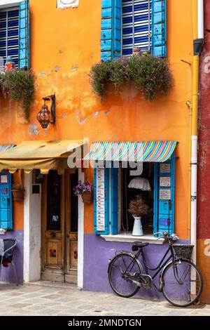 Ein junger Mann ruht vor einem bunt bemalten Laden auf Burano, durchsucht sein Handy, ein Fahrrad lehnt gegen die Wand Stockfoto