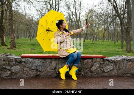 Glückliche lächelnde Seniorin in gelbem Regenmantel mit gelbem Regenschirm, die Selfie im Frühling-Regenpark macht. Ältere, schöne Frau mit Handy Stockfoto