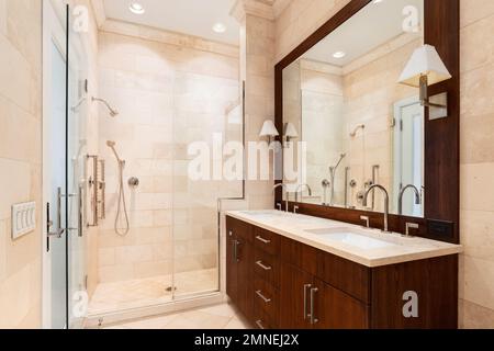 Ein luxuriöses Badezimmer mit dunklem Holzschrank und Spiegel, hellbraunen Fliesen an Wänden und Boden und einer begehbaren Dusche mit Glastür. Stockfoto