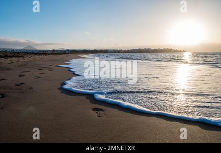 Strand von Lanzarote am Morgen mit Fußstapfen und einer Welle, die den goldenen Sand streichelt. Sonnenaufgang am Strand ohne Leute. Stockfoto