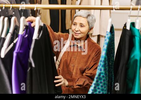 Vorderansicht einer lächelnden Seniorin, die in einem luxuriösen Boutiquen, Kopierraum, auf Kleiderständern herumschnüffelt Stockfoto