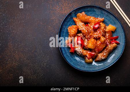 Traditionelles chinesisches Wok-Gericht, süßes und sauriges, frittiertes Hähnchen mit Gemüse, unter Rühren gebraten auf einem Teller mit Sesamsamen auf rustikalem dunklem Betonhintergrund Stockfoto