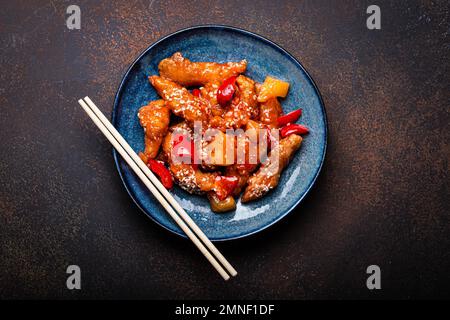 Traditionelles chinesisches Wok-Gericht, Schweiß und saure, frittierte Hähnchen mit Gemüse, gebraten auf einem Teller mit Sesamsamen auf rustikalem dunklem Betonstein Stockfoto