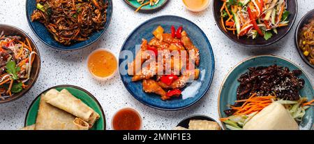 Set chinesischer Gerichte auf weißem Tisch: Süßes und saures Hähnchen, gebratene Frühlingsrollen, Nudeln, Reis, gedämpfte Brötchen mit bbq-glasiertem Schweinefleisch, asiatischer Stil Stockfoto