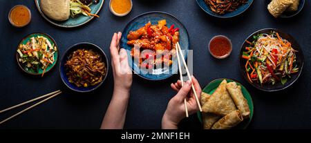 Set chinesischer Gerichte auf dem Tisch, weibliche Hände mit Stäbchen: Süßes und sauriges Hähnchen, gebratene Frühlingsrollen, Nudeln, Reis, gedämpfte Brötchen mit bbq Stockfoto