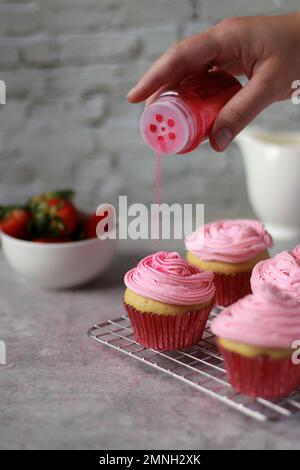 Hand stroht rosa Streusel auf Muffins mit Erdbeer Buttercreme Zuckerguss auf der Oberseite Stockfoto