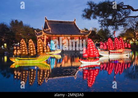 Beleuchtete chinesische Junks-Laternen und Friendship Hall-Pavillon am Dream Lake während der jährlichen Ausstellung Magic of Laternen im Chinesischen Garten in der Abenddämmerung. Stockfoto