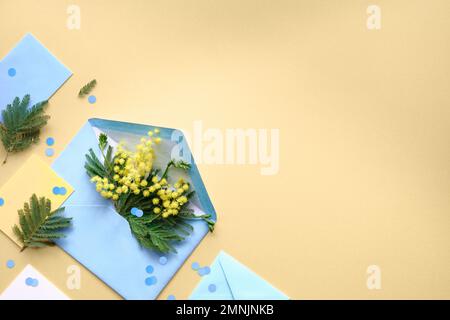 Frische Mimosenblüten in blauem Papierumschlag. Geometrisch flach liegend, Draufsicht mit Blumen und Grußkarten auf gelbem Hintergrund, Kopierbereich Stockfoto