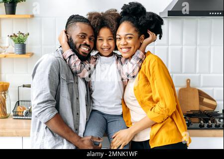 Familienporträt. Afroamerikanische glückliche Familie, Dad, mom und ihre süße Vorschultochter, die sich in einer Küche zu Hause umarmen, in die Kamera schauen, lächeln, glücklich zusammen sein, Mädchen umarmt ihre Eltern Stockfoto