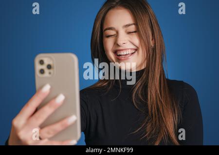 Porträt einer charmanten jungen Frau, die sanft an der Hand zieht, mit Smartphone in der Nähe des Gesichts, um ein Foto auf blauem Hintergrund zu machen. Stockfoto