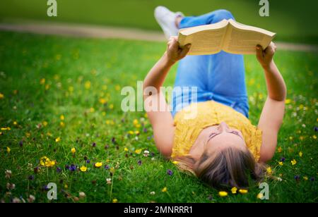 Verirren Sie sich in Literatur. Eine sorgenfreie junge Frau, die sich mit einem Buch auf einem Wiesenfeld entspannt. Stockfoto