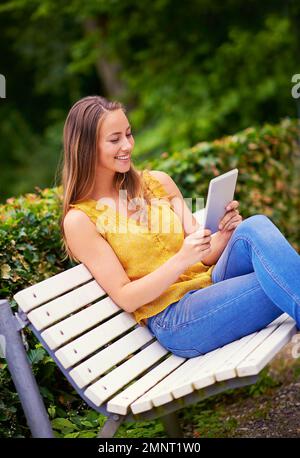 Ich melde mich bei ihren Online-Freunden. Eine junge Frau, die ein digitales Tablet auf einer Parkbank benutzt. Stockfoto