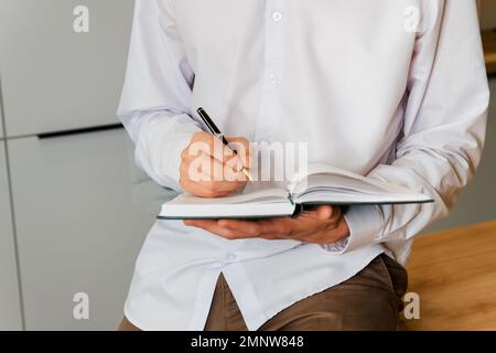 Ein erwachsener Mann sitzt auf einem weichen Stuhl und schreibt Notizen von Hand in ein Notizbuch Stockfoto