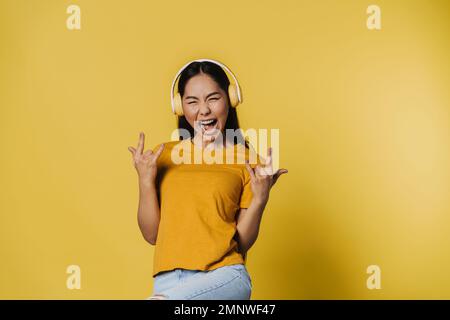Junge asiatische Frau in Kopfhörern schreit, während sie sich isoliert über gelben Hintergrund bewegt Stockfoto
