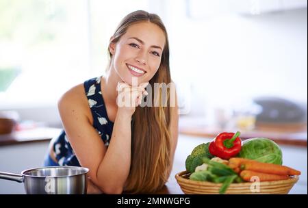 Lieben Sie sich genug, um einen gesunden Lebensstil zu führen. Porträt einer jungen Frau, die sich auf ihren Küchentisch lehnt, mit einem Topf und Gemüse neben ihr. Stockfoto