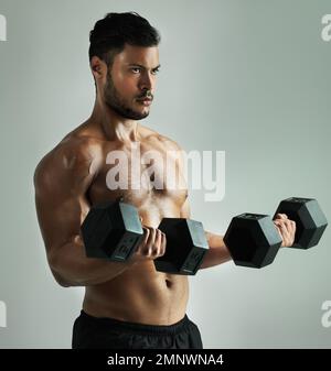 Trainiere verrückt oder bleib gleich. Studioaufnahme eines jungen Mannes, der mit Kurzhanteln trainiert, vor grauem Hintergrund. Stockfoto