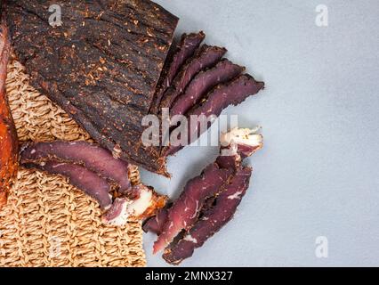 Frisch geschnittene südafrikanische Biltong mit selektivem Fokus auf grauer Oberfläche mit gewebter Tischmatte Stockfoto