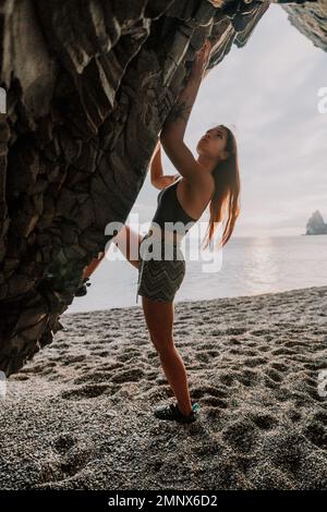 Sportlerin Klettert Auf Den Felsen. Junge Frau mit schlankem Körper klettert in einer vulkanischen Basalthöhle mit wunderschönem Blick auf das Meer. Das Sportlermädchen trainiert Stockfoto