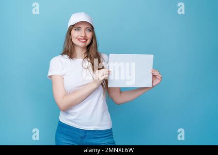 Wunderschöne junge Frau mit leerem weißem Pappbrett, Ihr Text hier Konzept, weißliches Mädchen in weißem T-Shirt und Mütze für Modell isoliert über einem farbigen, blauen Hintergrund Studio Stockfoto