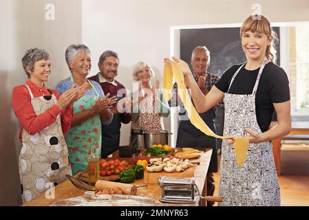 Das Leben ist eine Kombination aus Magie und Pasta. Porträt einer Frau, die ihre frisch zubereitete Pasta mit ihren Schülern im Hintergrund hielt. Stockfoto