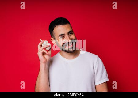 Ein junger bärtiger Mann in weißem Hemd hält Knoblauch mit einem Lächeln, isoliert auf rotem Hintergrund Stockfoto