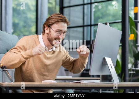 Ein junger Mann im Amt feiert den Sieg und erfolgreiche Erfolge, ein Geschäftsmann, der mit Triumph-Geste Händchen hält und gute Nachrichten aus dem Computer liest, ein blonder Programmierer in lässiger Kleidung. Stockfoto