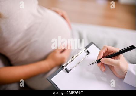 Nahaufnahme einer professionellen Ärztin überprüft ihre schwangere Patientin, während sie eine Krankenakte auf ein medizinisches Klemmbrett schreibt. Stockfoto