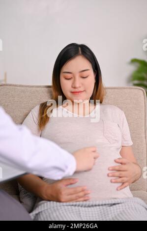 Glückliche junge asiatische Schwangere wird von ihrem Arzt in der Klinik untersucht. Ein Arzt, der den Herzschlag des Babys mit einem Stethoskop hört. Stockfoto