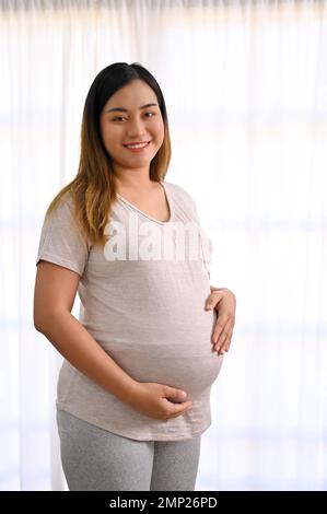 Das Porträt einer glücklichen und schönen jungen asiatischen schwangeren Frau in lässiger, bequemer Kleidung steht am Fenster und berührt ihren Bauch mit Liebe. Stockfoto