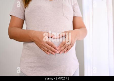 Gekürzte Aufnahme einer schwangeren Frau in bequemen Kleidern, die ihre Herzhände auf ihrem großen Bauch unterschreiben lässt. Liebe, Erwartung, Erziehung, Mutterschaftskonzept Stockfoto