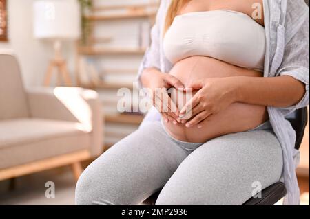 Glückliche schwangere Frau, die ein Herz-Hand-Zeichen auf ihrem Bauch macht. Mutterschaft, Schwangerschaft, Liebe, Erwartung. Freigestellte Aufnahme Stockfoto