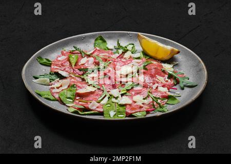 Carpaccio aus Marmorfleisch mit Rucola und Mangold, Zitrone und Parmesankäse auf dunklem Hintergrund Stockfoto