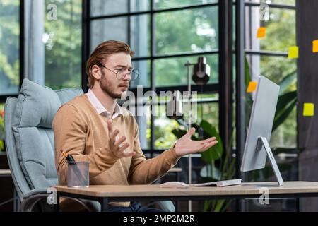 Frustrierter und verärgerter Geschäftsmann, der online Nachrichten auf dem Computermonitor liest, junger blonder Mann, der im Büro in lässiger Kleidung traurig ist und mit den Ergebnissen nicht zufrieden ist. Stockfoto